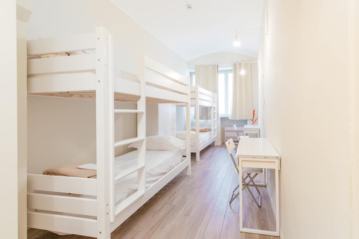 A bunk bed room with a desk and chair, wooden flooring, and a window with curtains.