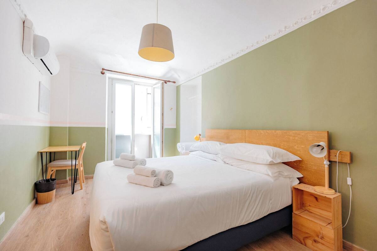 A neatly arranged bedroom with a bed, bedside table, lamp, and a small desk with a chair.