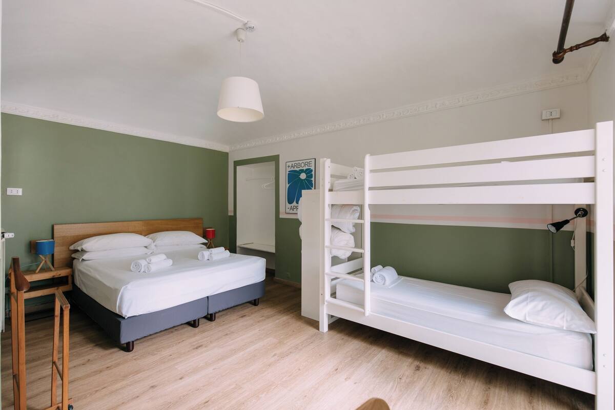 A room with a bunk bed, a single bed, a bedside table with a lamp, and a wall-mounted shelf.