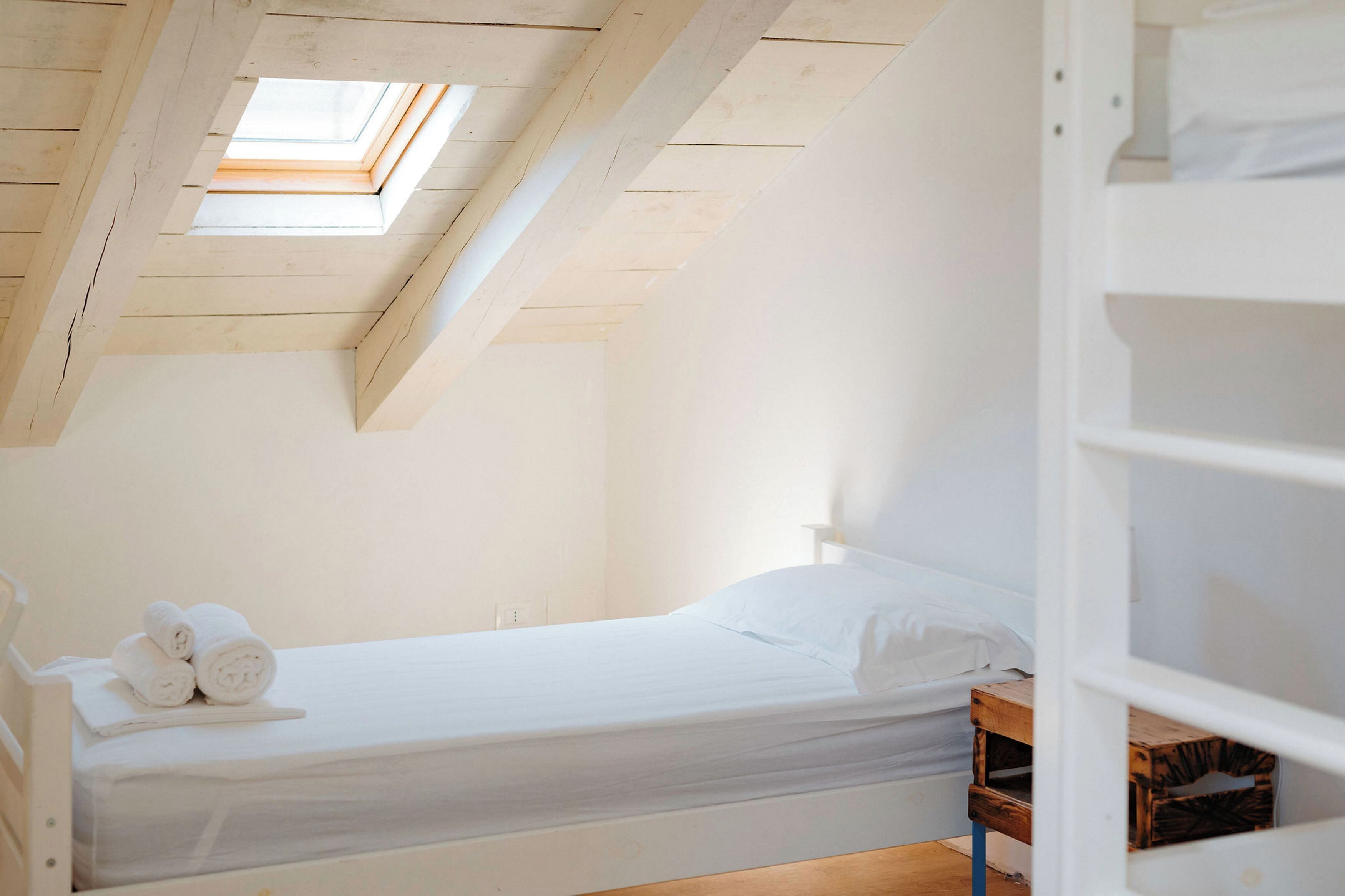 Une chambre avec un puits de lumière, un lit recouvert de draps blancs et une table de chevet en bois.