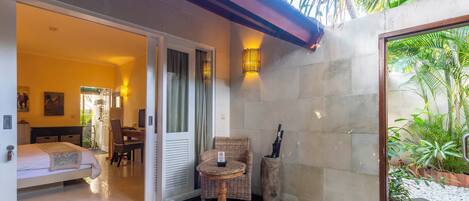 Una habitación con cama, una mesa de comedor y una silla de mimbre. Hay una pequeña mesa con una silla afuera de la habitación.