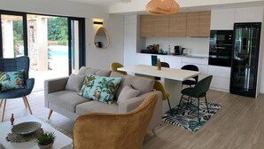 Una sala moderna con un sofá, sillones y un comedor con mesa y sillas. Cuenta con una cocina con armarios de madera y un refrigerador.