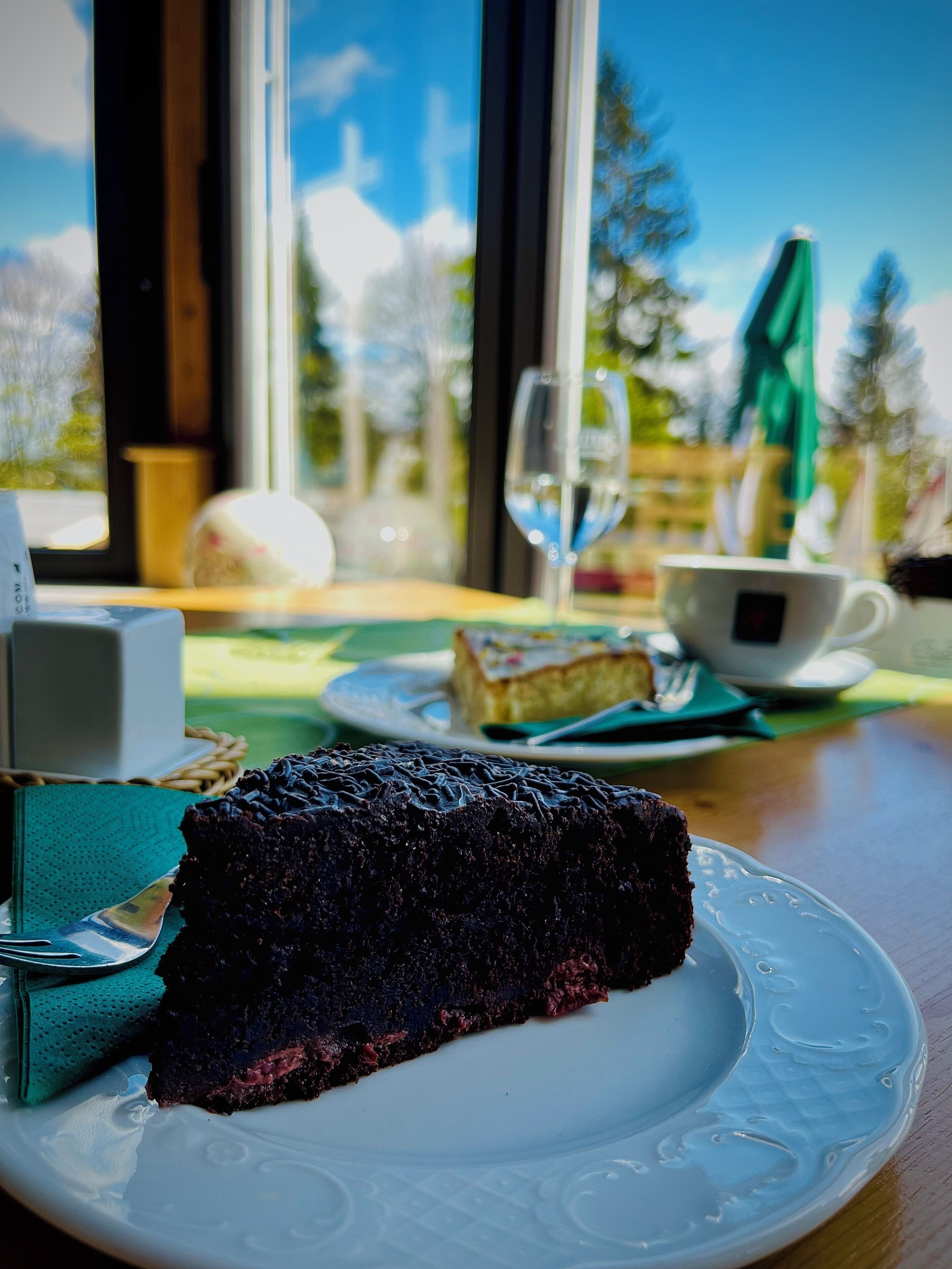 Ein Stück Schokoladenkuchen auf einem weißen Teller, ein Glas Weißwein und eine Tasse Tee auf einem Tisch mit Blick auf Bäume und einen blauen Himmel durch ein Fenster.