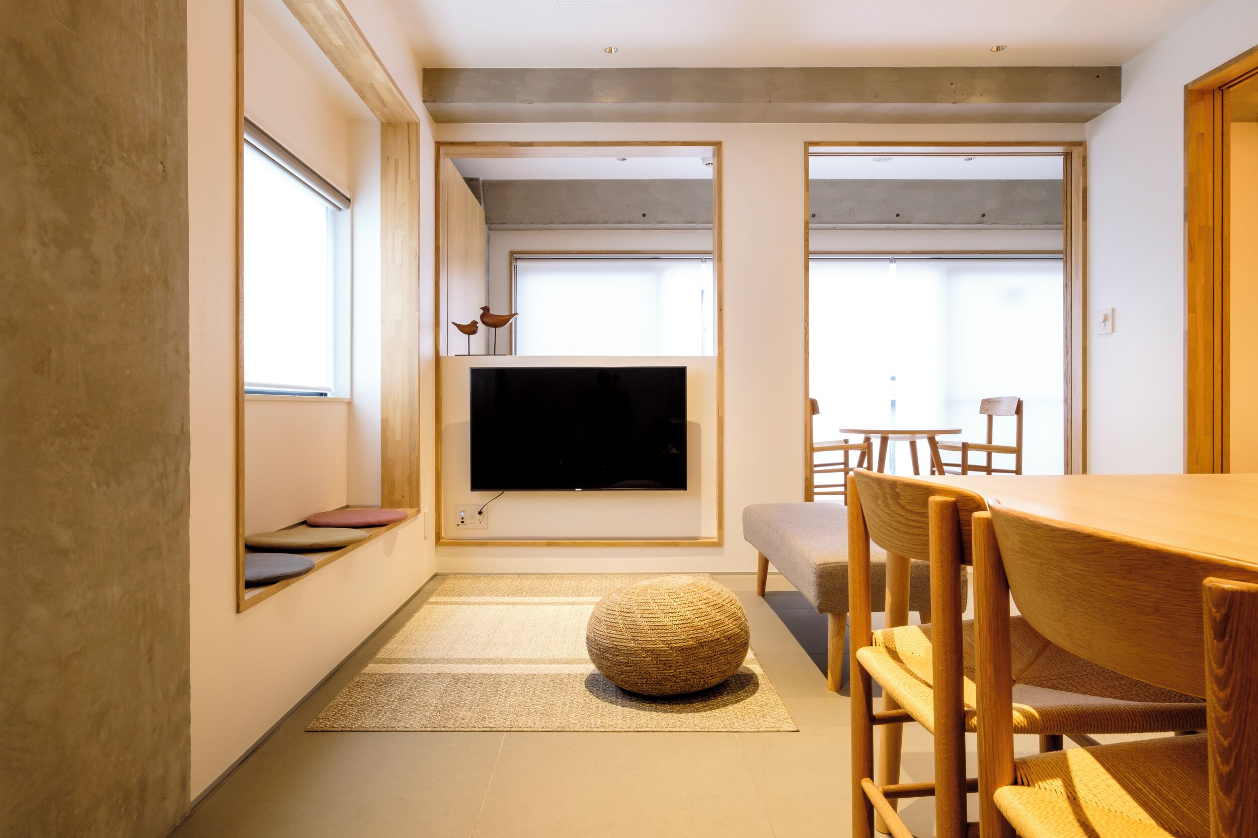 Una sala moderna con un televisor de pantalla plana grande montado en la pared, una mesa de comedor de madera con sillas y un otoman tejido.