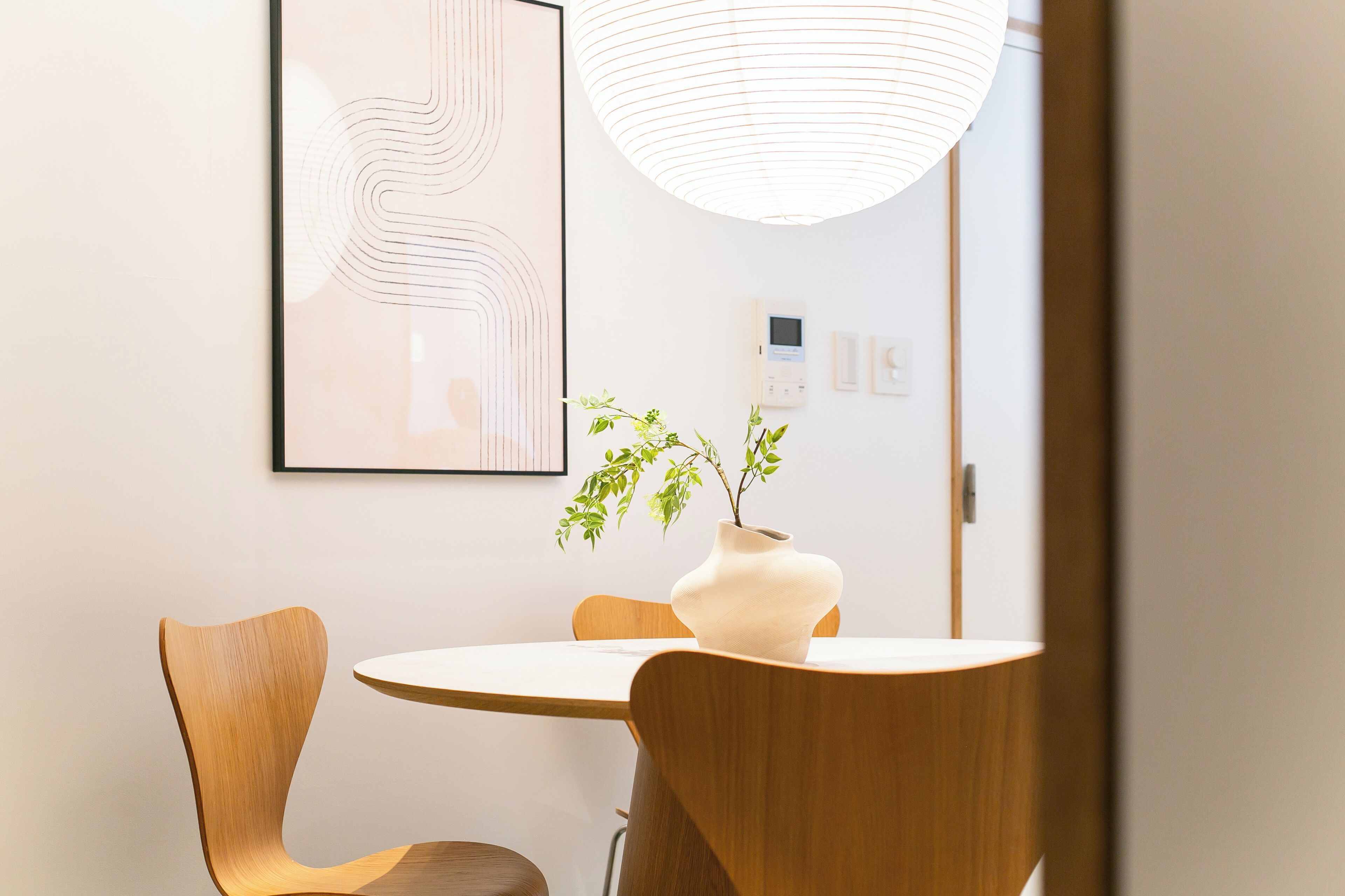 A modern dining area with a round table, wooden chairs, a vase with a plant, a pendant light, and a framed abstract artwork on the wall.