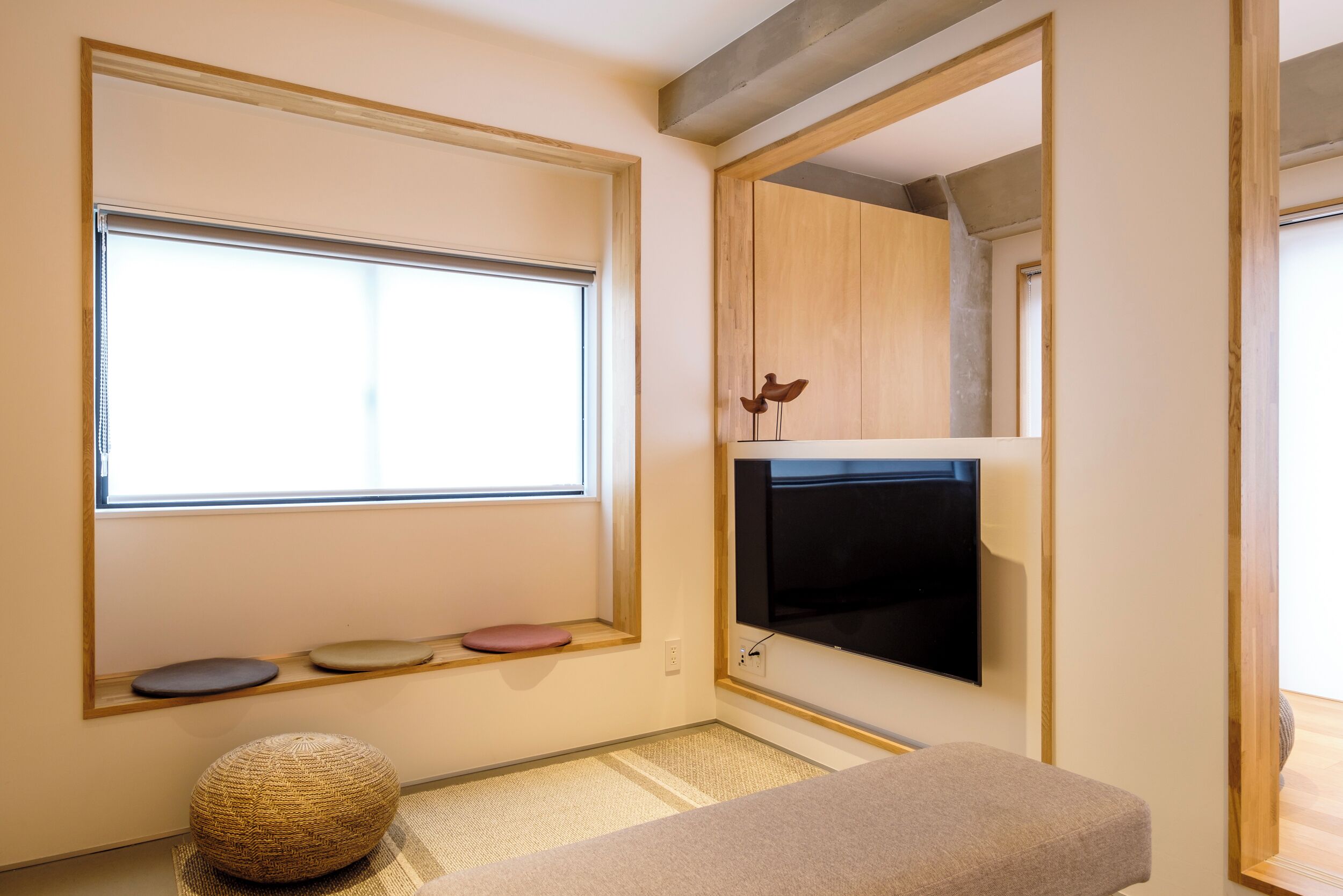 A room with a large window, a flat-screen TV mounted on the wall, a small shelf with decorative items, and a woven pouf on the floor.