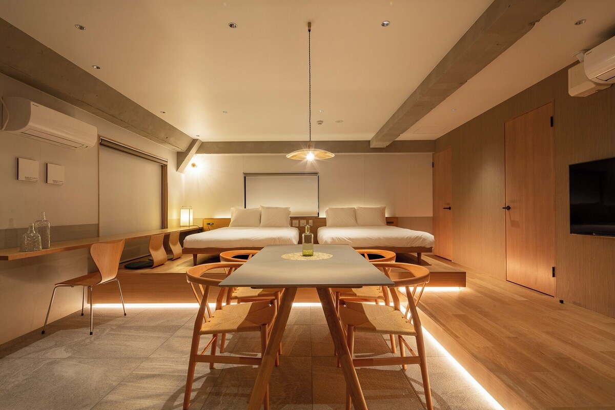 A modern hotel room with a central dining table, two beds, a TV, and a wooden wardrobe.