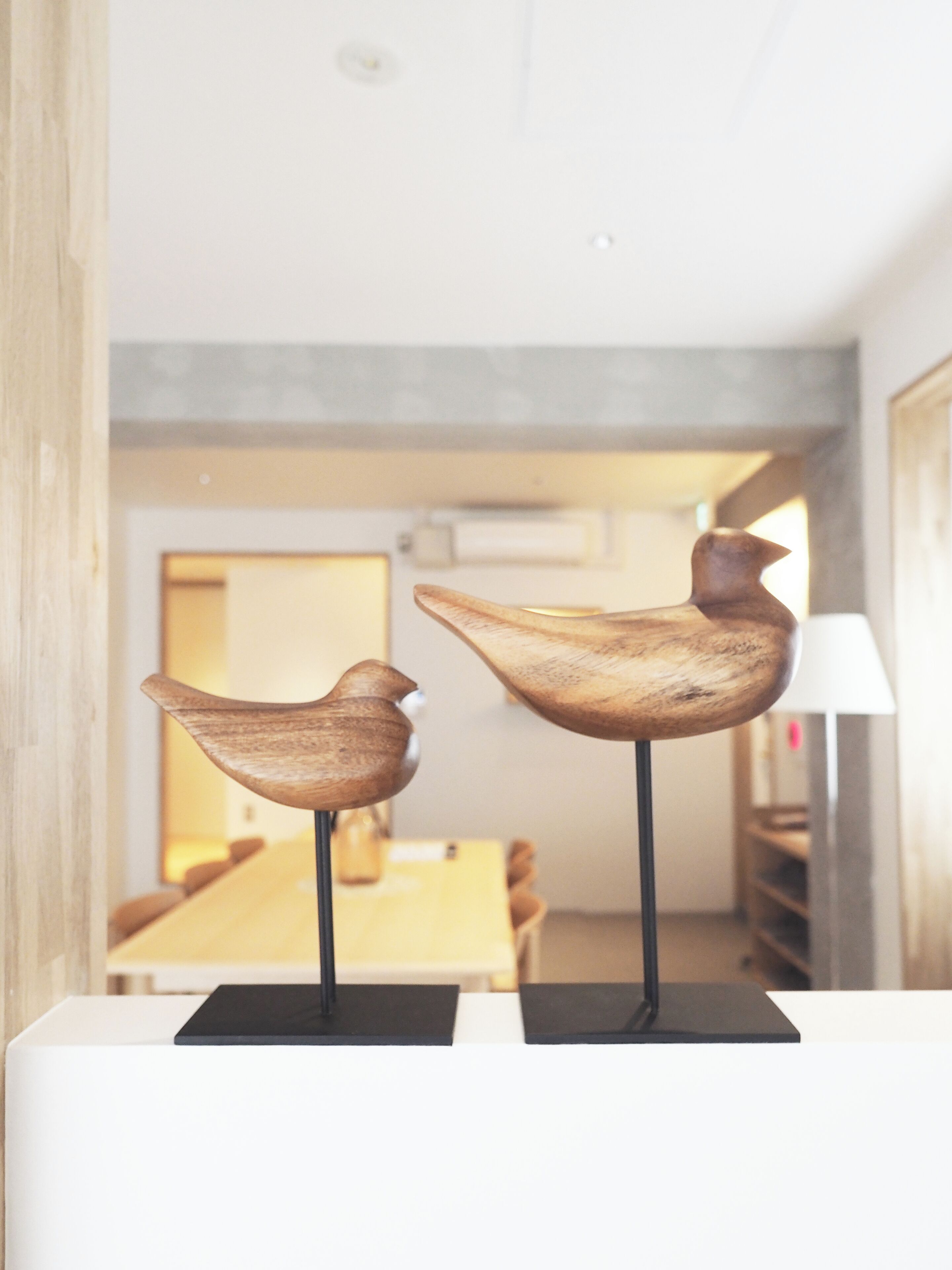 Two wooden bird sculptures on black pedestals in a room with a wooden table and chairs in the background.