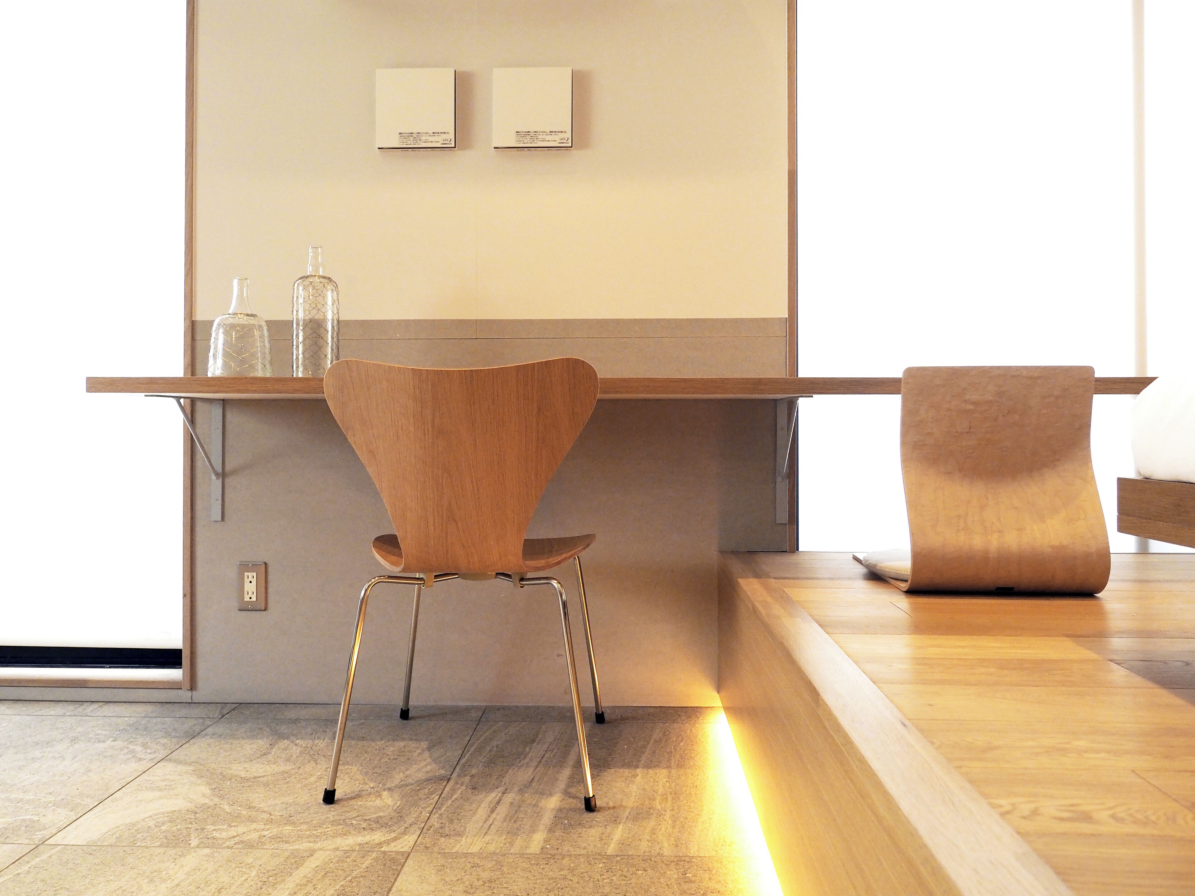 Un espace de travail moderne et minimaliste, avec un bureau en bois, une chaise et un banc. Deux objets encadrés sont accrochés au mur et une bouteille est posée sur le bureau.