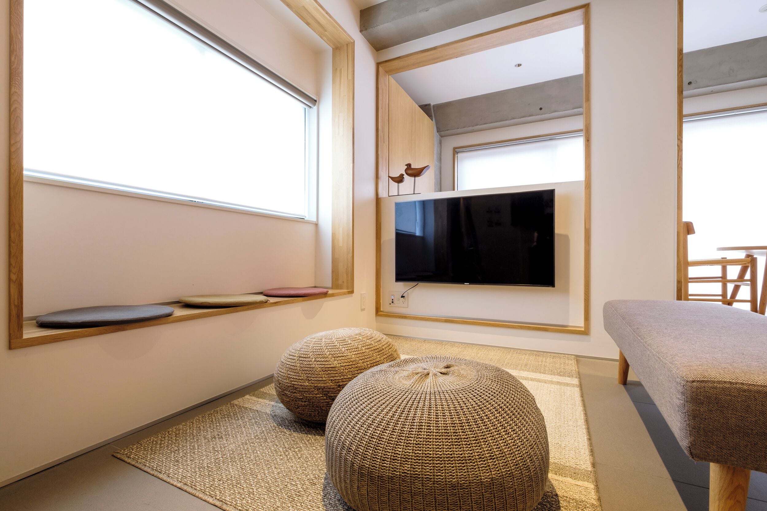 A modern living room with a large flat-screen TV mounted on the wall, two round woven ottomans, a grey sofa, and a wooden bench.