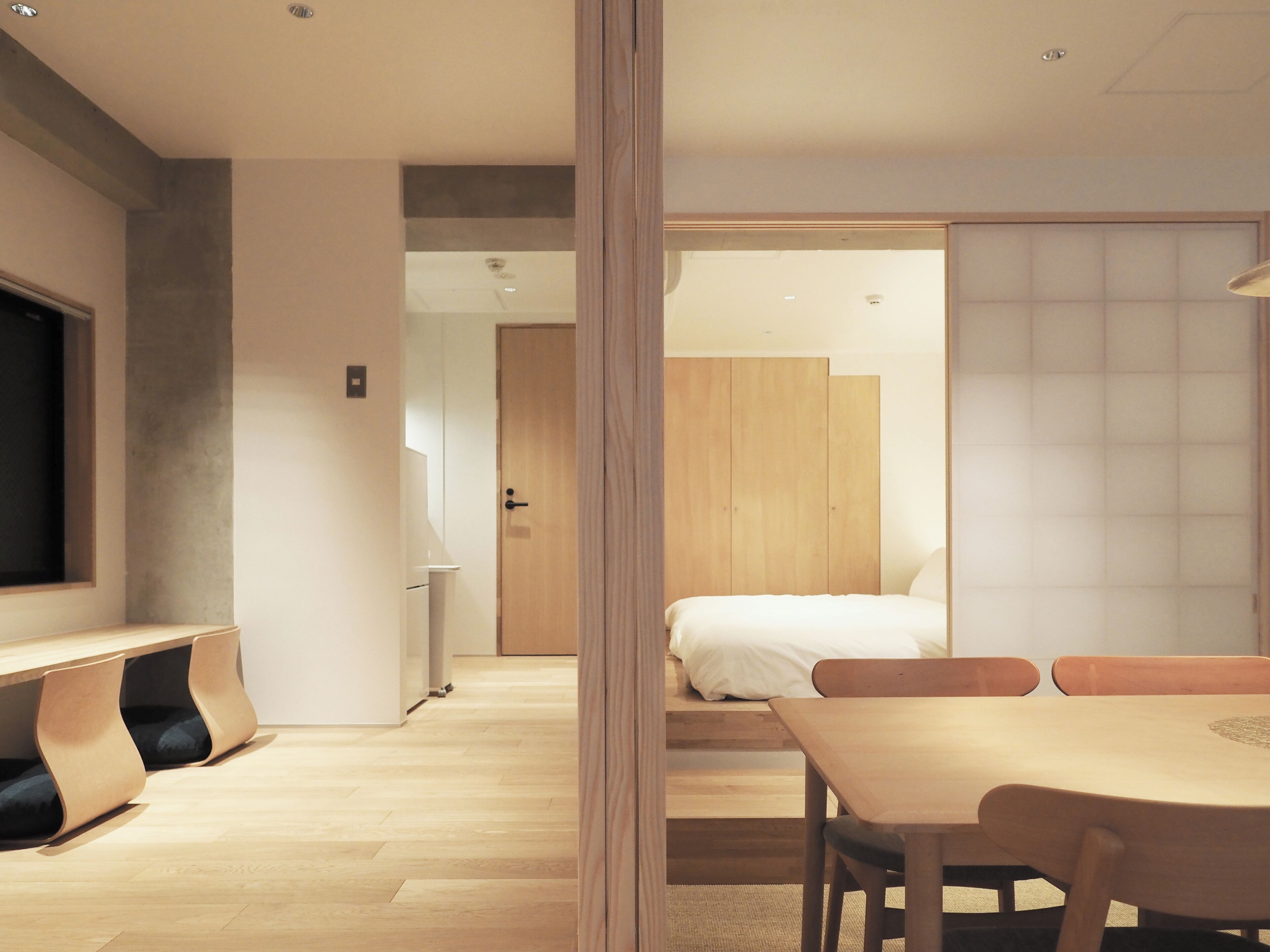 A modern hotel room with a wooden floor, a bed with white bedding, a wooden desk, and a television on the left.