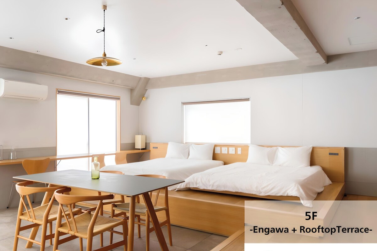 A modern hotel room with a bed, a dining table with chairs, a small kitchen area, and a large window.