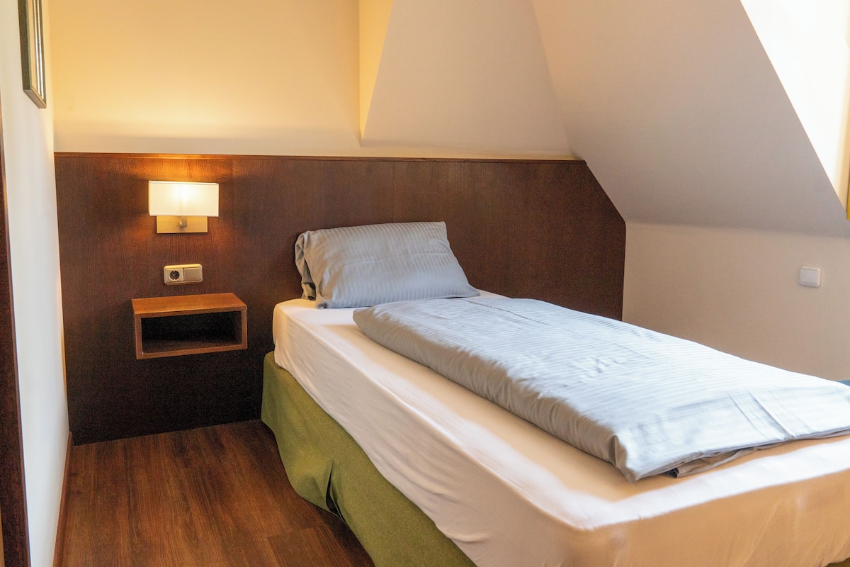 A small, neatly made bed in a room with a sloped ceiling, a bedside table with a lamp, and a wall-mounted light fixture.