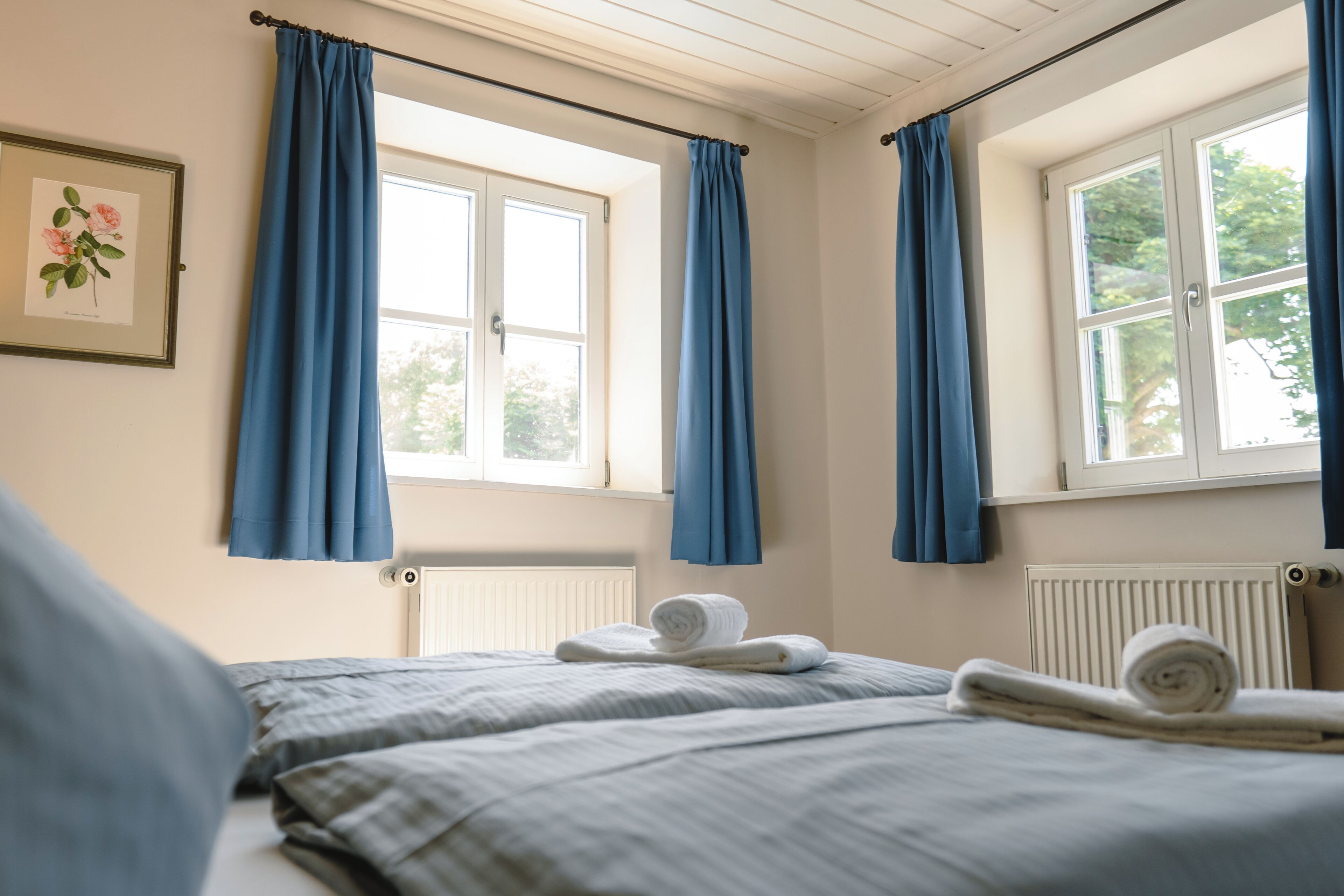 A bedroom with two beds, blue curtains, a framed botanical print, and a rolled towel on the bed.