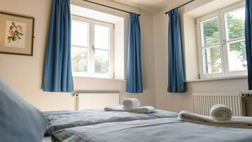A bedroom with two beds, blue curtains, a framed botanical print, and a rolled towel on the bed.