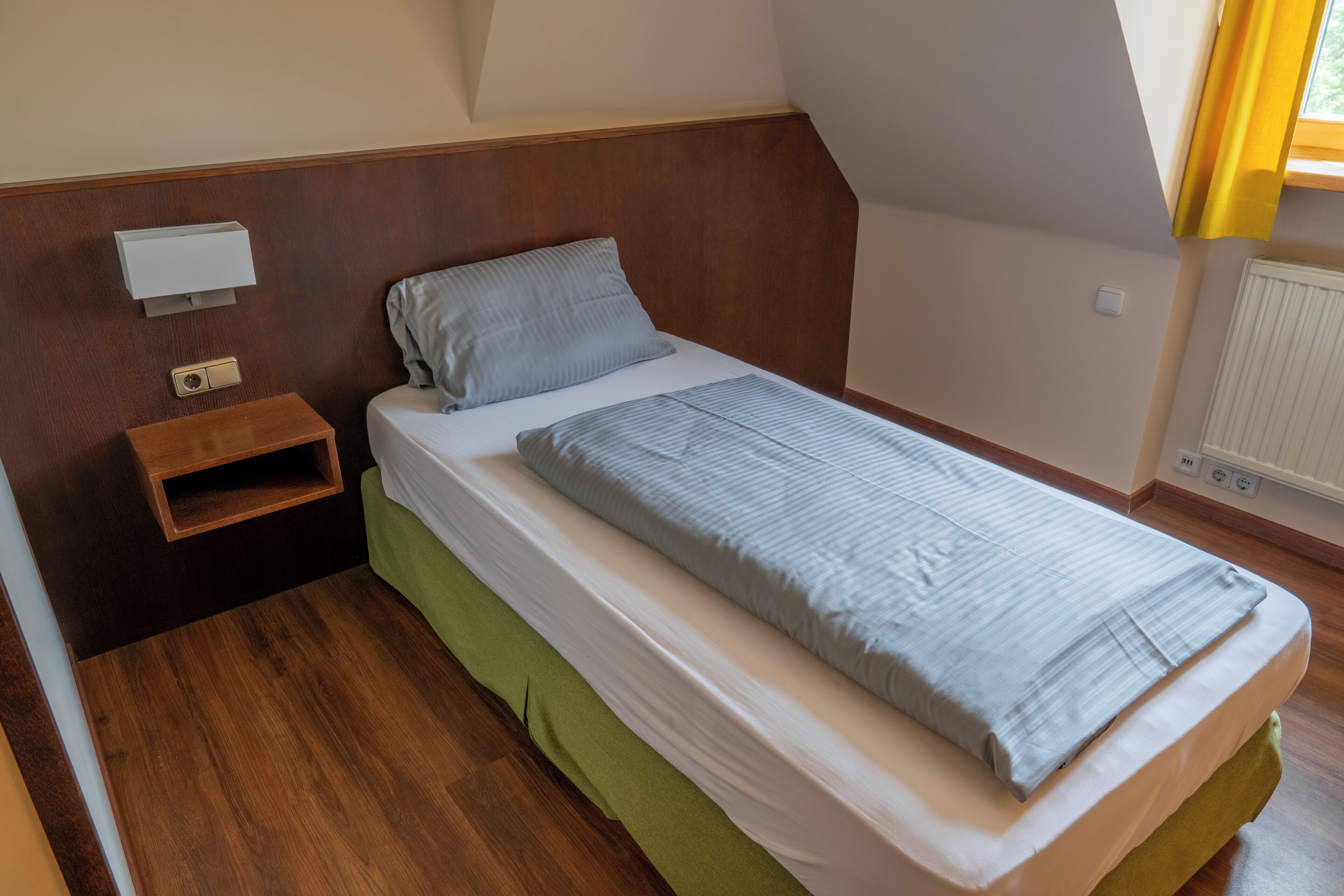 A small, neatly arranged bedroom with a single bed, a nightstand, a wall-mounted light, and a radiator.
