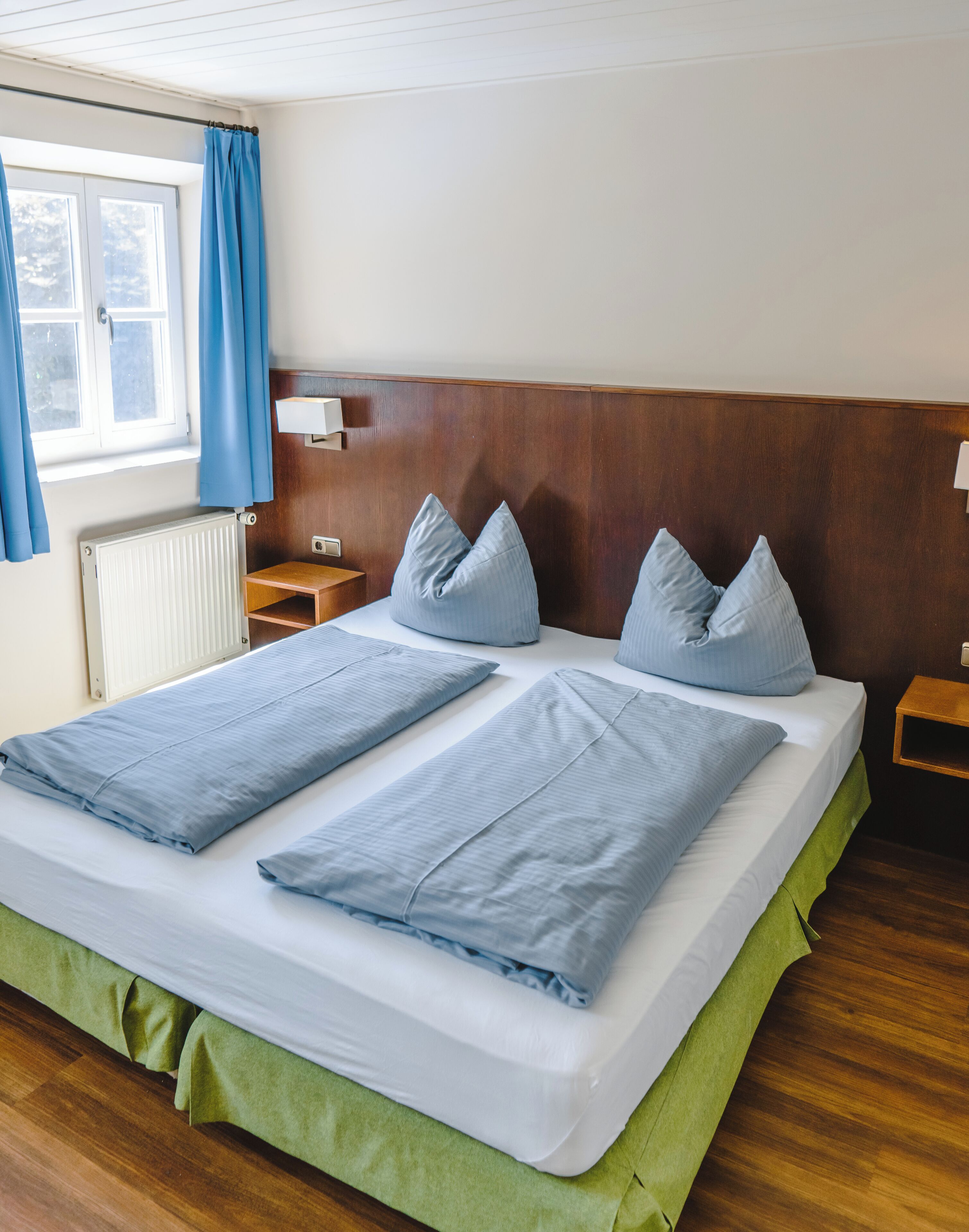 A double bed with green bed skirt, two nightstands, a wooden headboard, a window with blue curtains, and a white radiator.