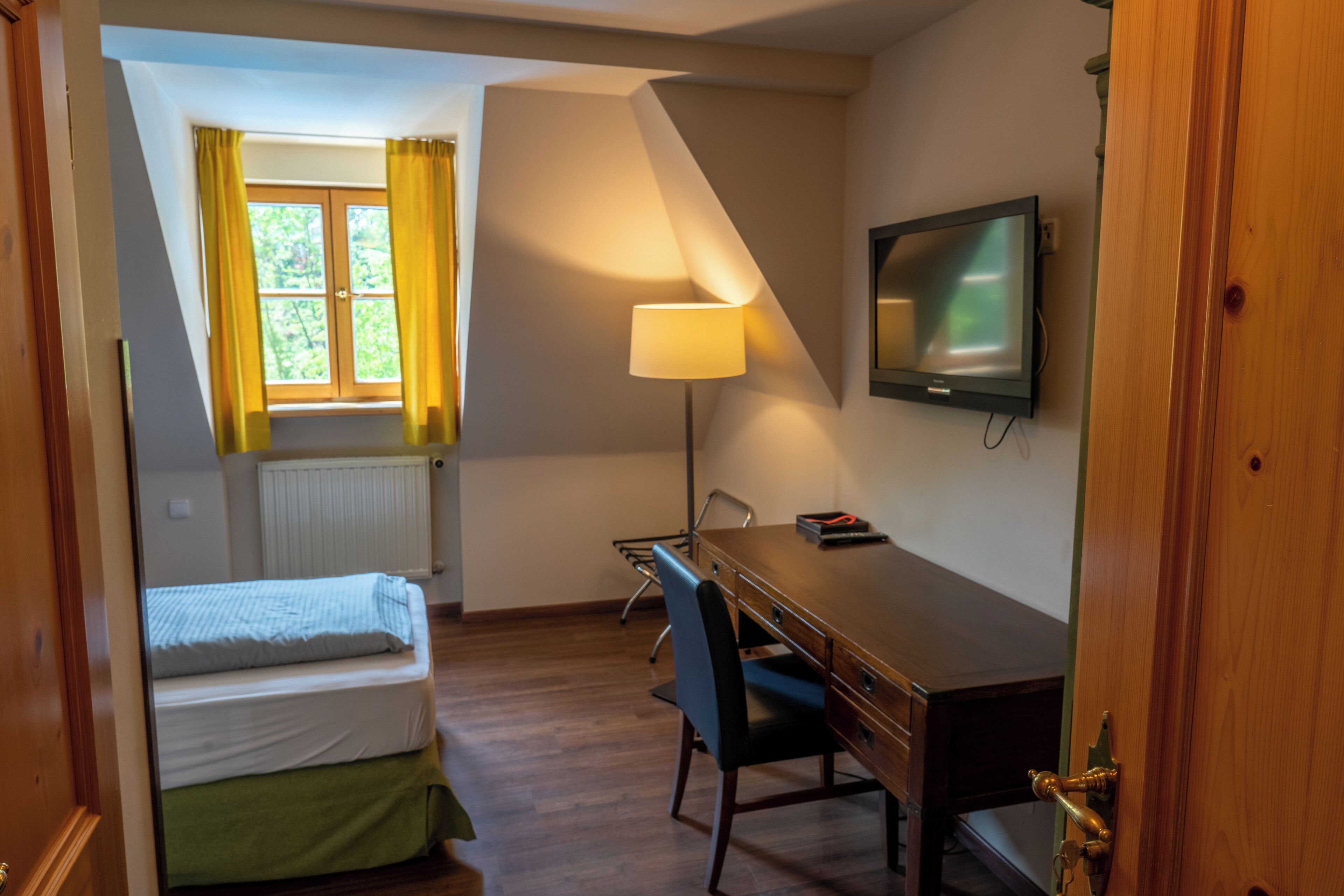 Ett hotellrum med en säng, ett skrivbord, en stol, en lampa, en tv och ett fönster med gardiner.