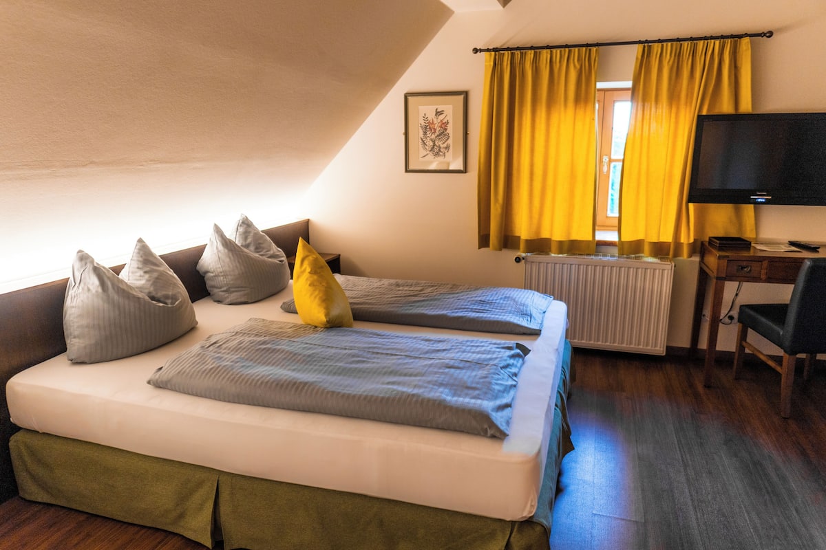 A room with two beds, a desk, a chair, a TV, and a window with yellow curtains.
