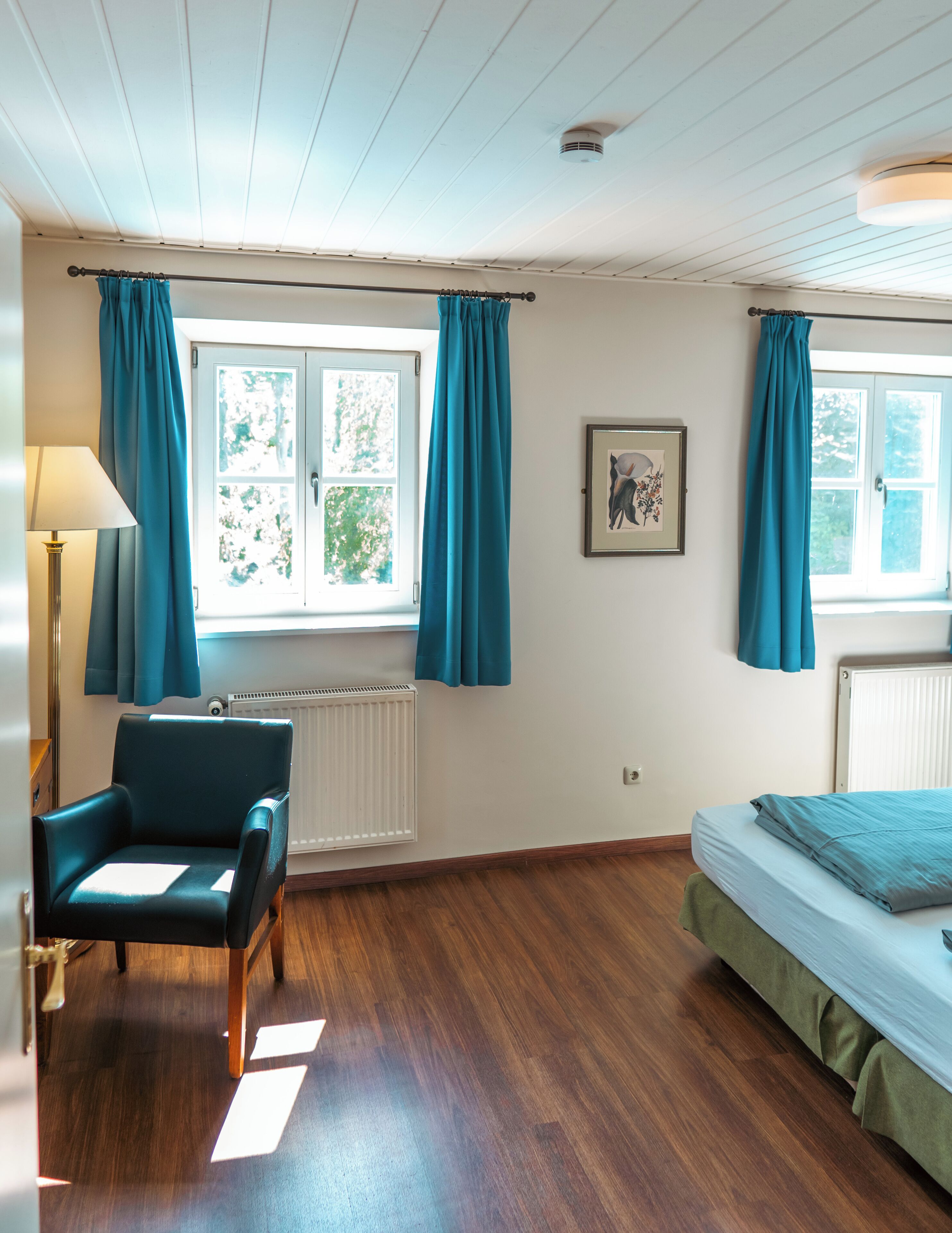 A room with a wooden floor, a chair, a bed, a radiator, and a window with curtains.