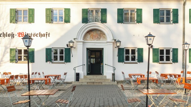 A building with a central entrance, multiple windows with green shutters, outdoor seating with tables and chairs, and a paved ground. - Noon Mirch Hotel Restaurant (Eching)