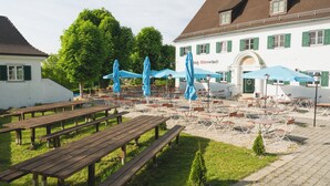 An outdoor seating area with wooden tables and blue umbrellas, surrounded by a white building with green shutters. - Noon Mirch Hotel Restaurant (Eching)