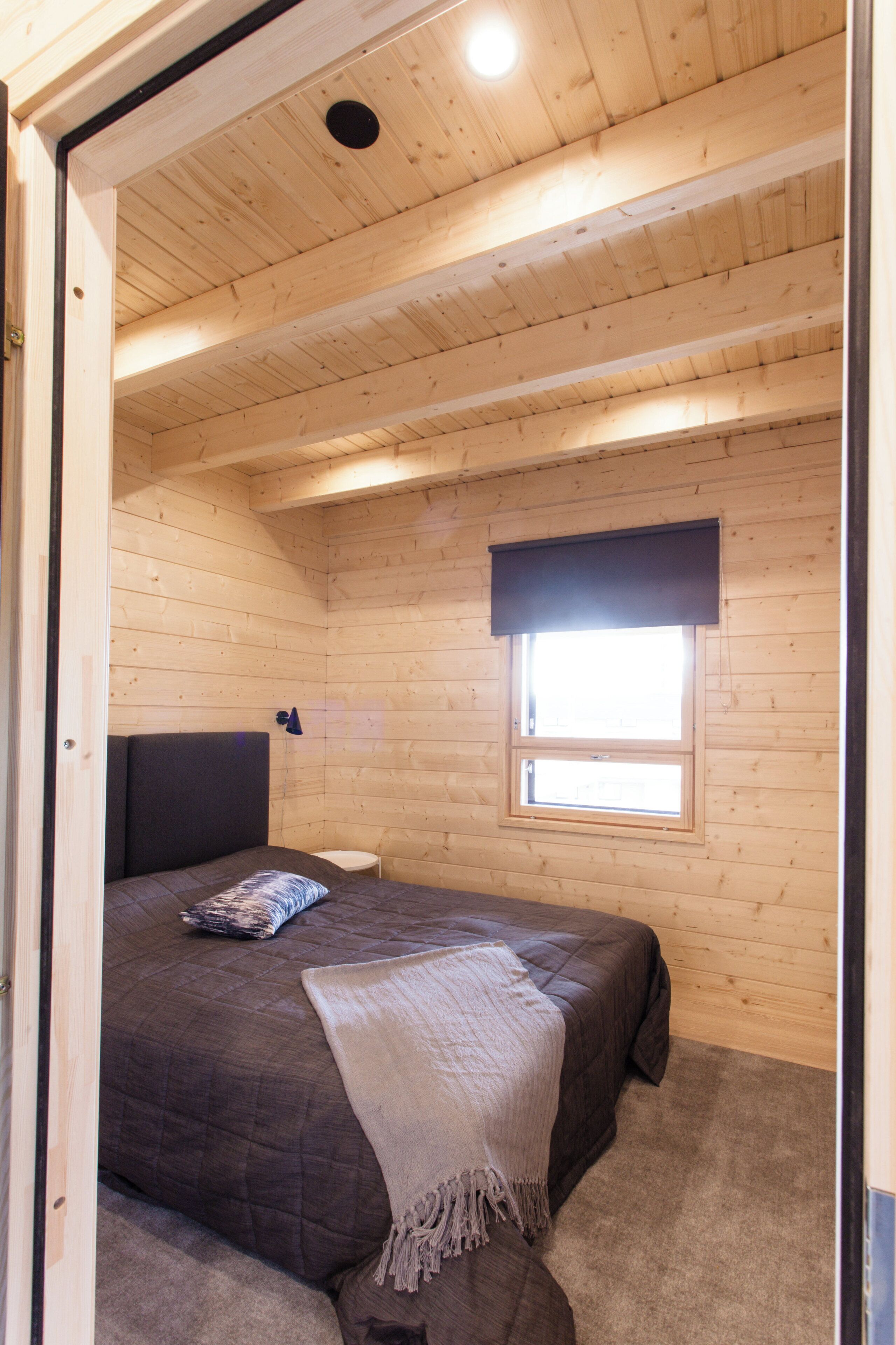 A wooden cabin bedroom with a bed, a window with a dark blind, and a ceiling light.