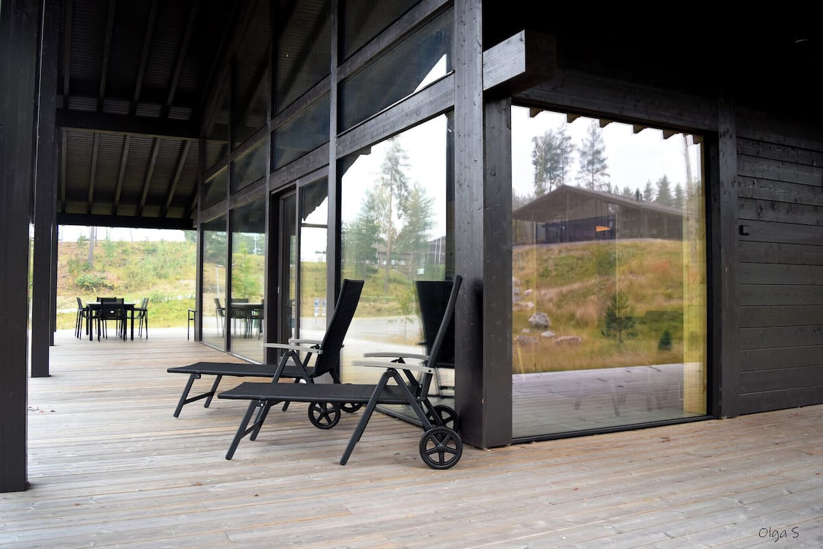A covered outdoor area with a wooden deck, large windows, and a view of a building and landscape.