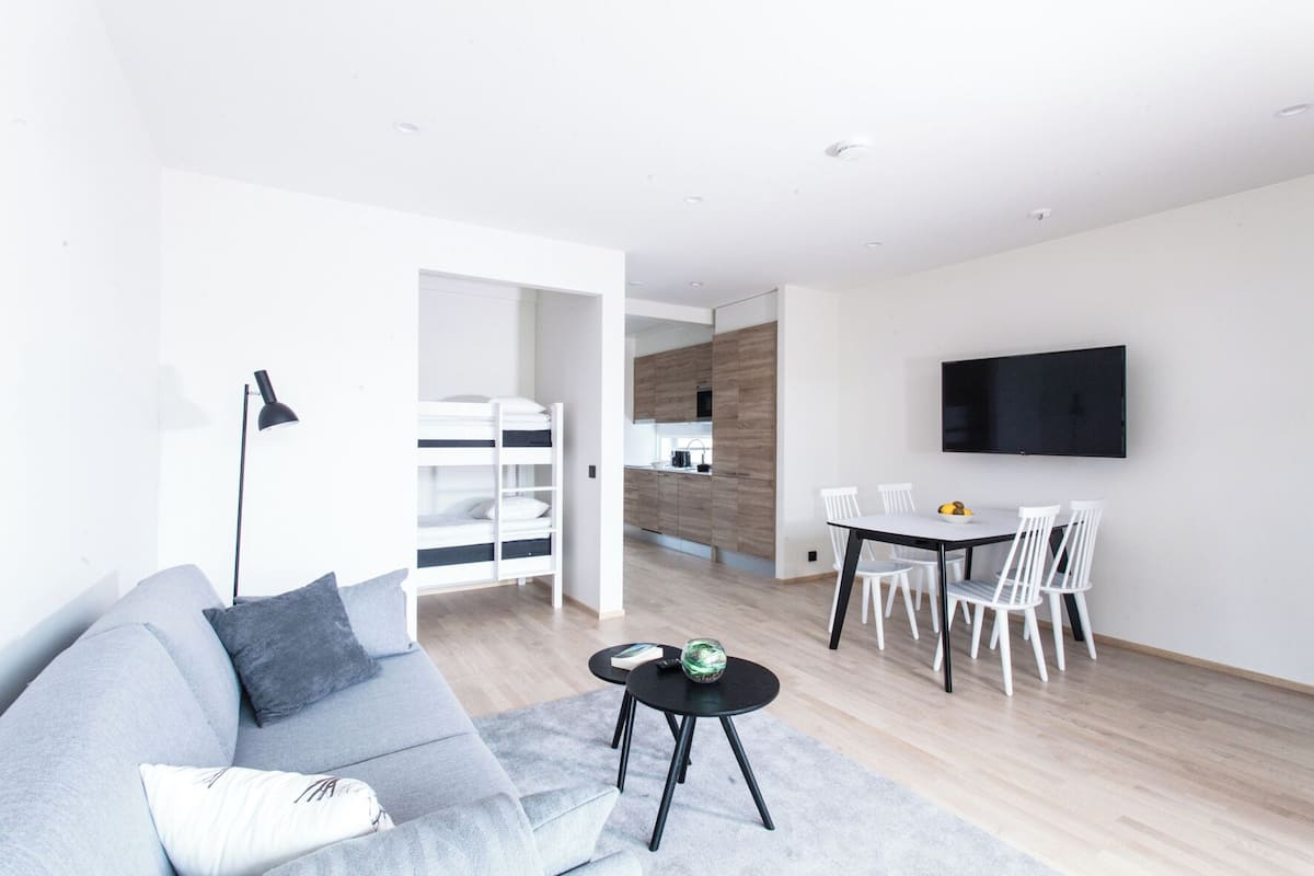 A modern living room with a grey sofa, a black coffee table, and a dining area with white chairs and a TV mounted on the wall.