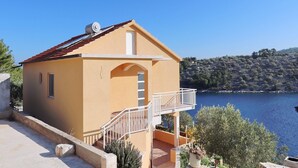 A small, single-story building with a balcony, a red-tiled roof, and a view of a body of water and hills in the background. - Peaceful Holiday House Senija (Vela Luka)