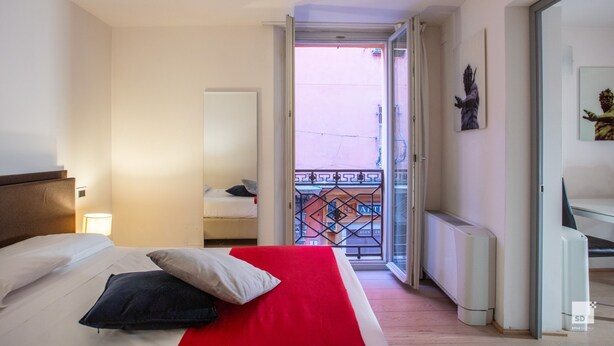 A hotel room with a bed, a nightstand, a lamp, a radiator, and a balcony with a decorative railing.