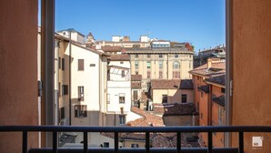 A view from a balcony overlooking a narrow street with traditional buildings, a clear blue sky, and a metal railing in the foreground.