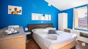 A modern hotel room with a large bed, a bedside table with a lamp, a dresser, a window with a view of buildings, and a blue wall with a framed picture.