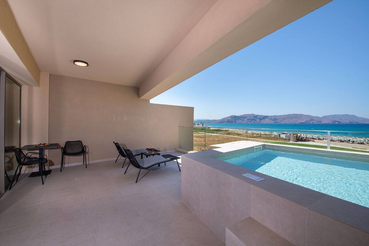 A balcony with a pool, chairs, and a table, overlooking a beach and mountains.