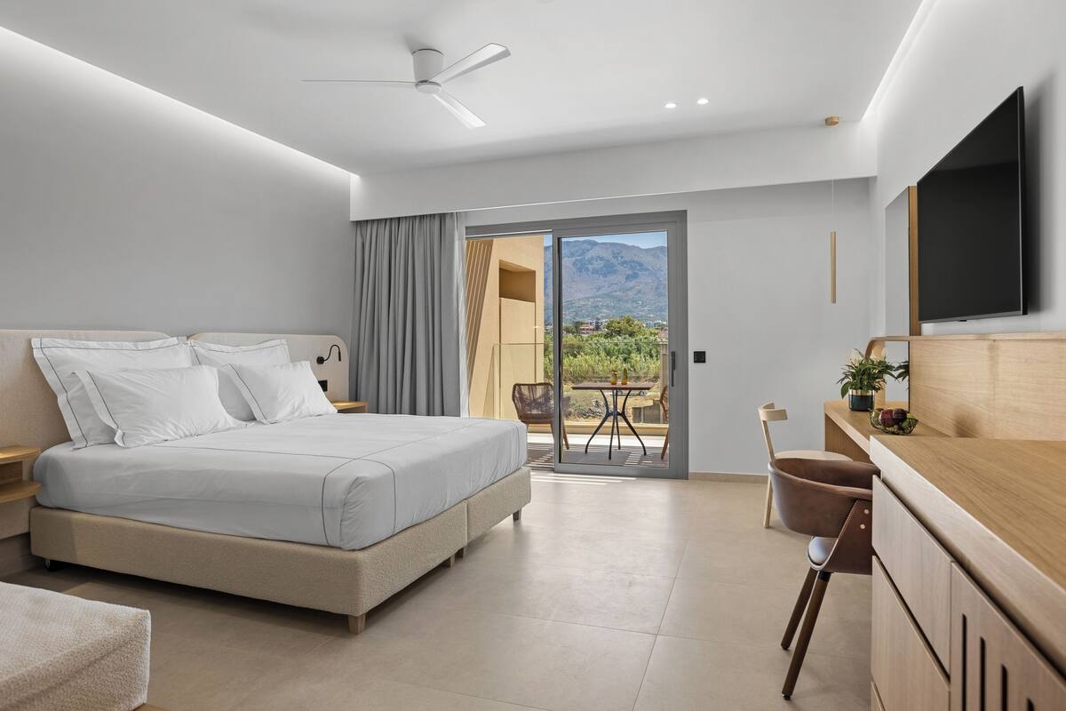 A modern hotel room with a large bed, a desk, a chair, a TV, and a balcony with a mountain view.