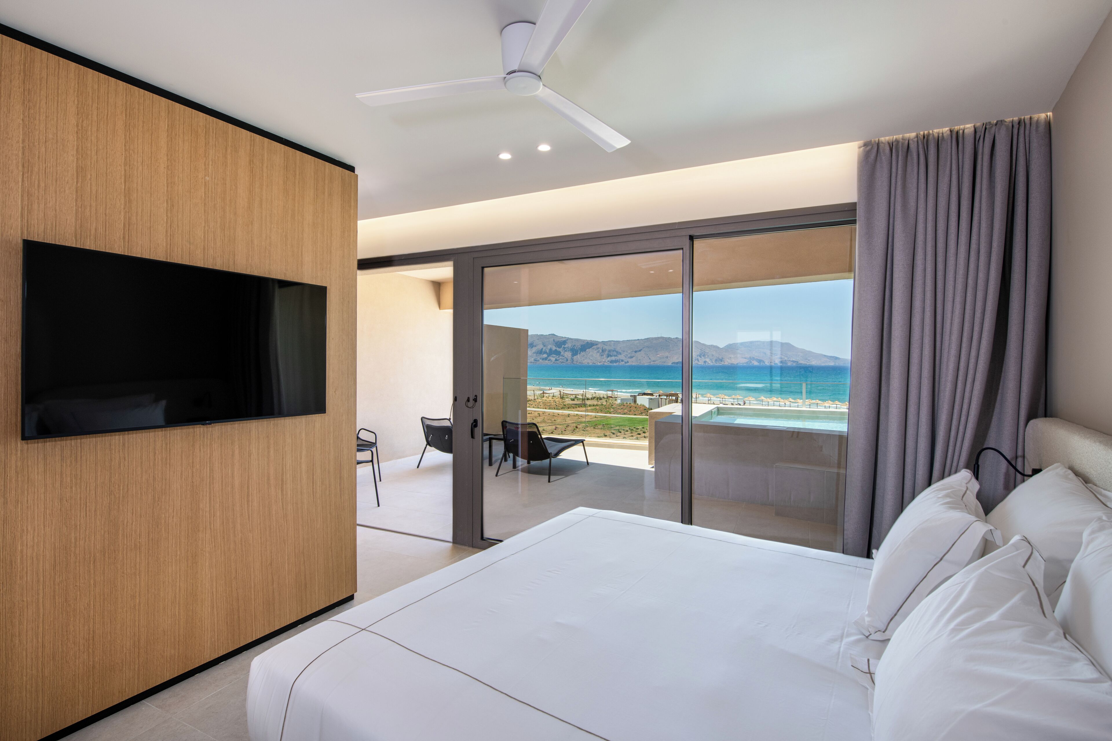 Una habitación de hotel moderna con una cama grande, un televisor de pantalla plana, un ventilador de techo y un balcón con vista a la playa y el océano.