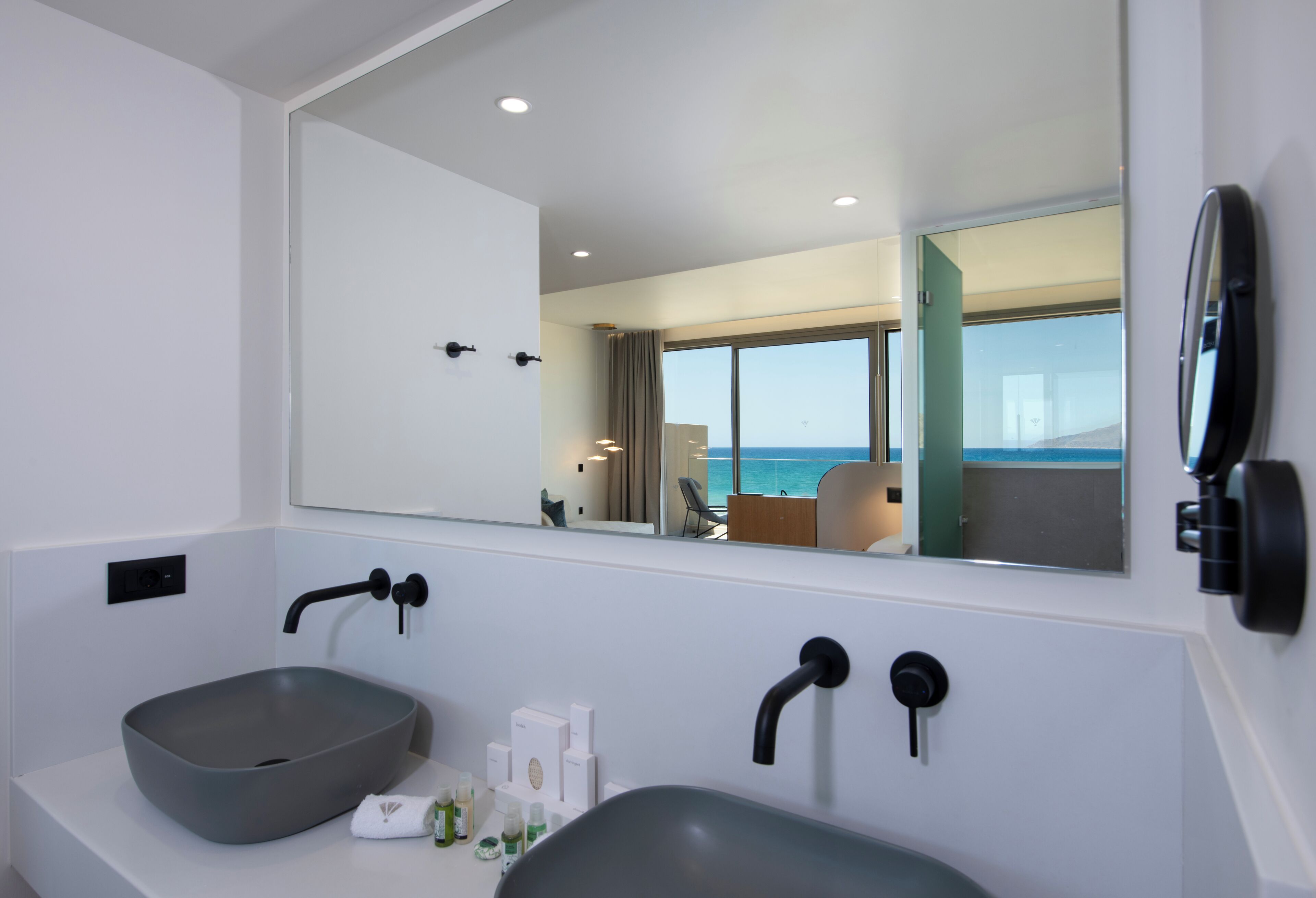 Un baño moderno con un espejo grande, dos lavamanos y vistas al mar a través de amplias ventanas.