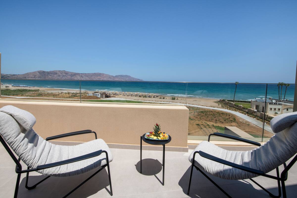 A balcony with two chairs and a small table with fruit, overlooking a beach and ocean.