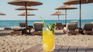 Een glas geel drank op een houten tafel, met strandparasols en ligstoelen op de achtergrond.