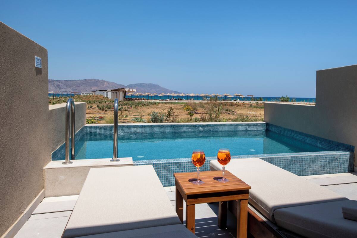 A pool with a view of the beach and mountains. There are lounge chairs and a table with two glasses.