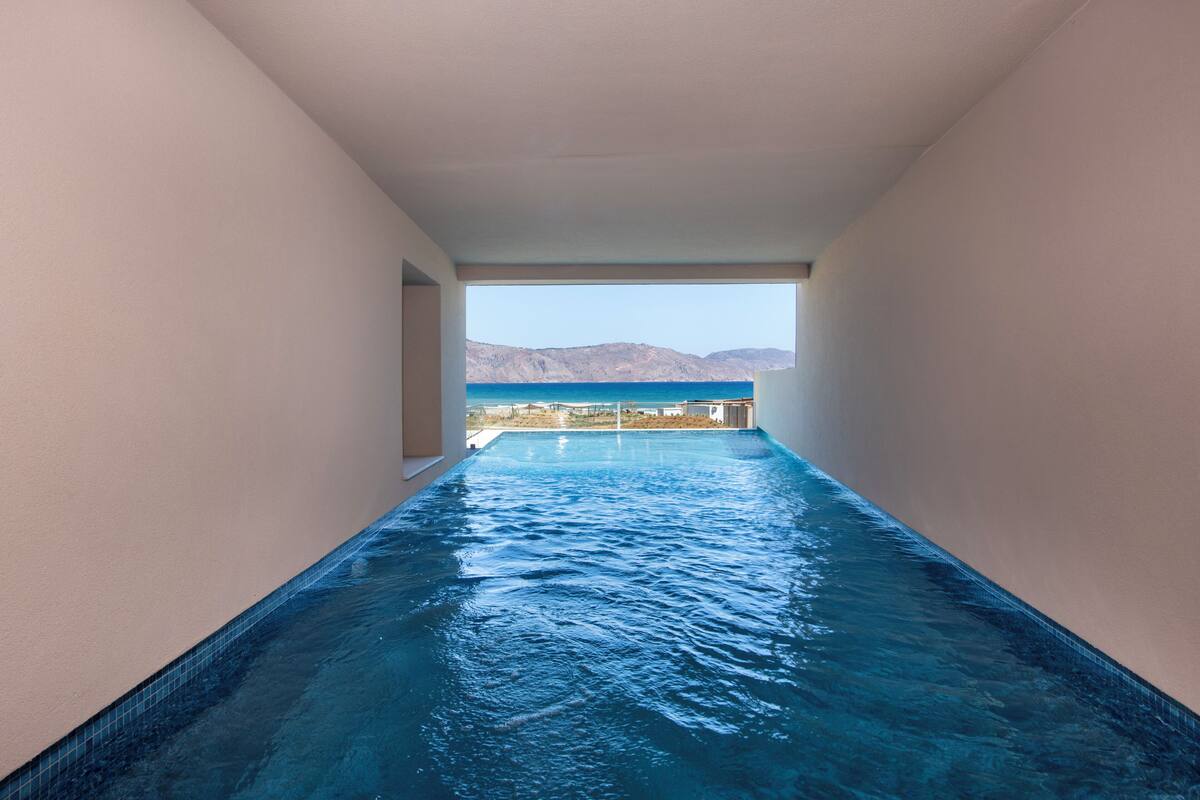 A narrow indoor swimming pool with clear blue water, leading to a balcony with a view of mountains and the sea.