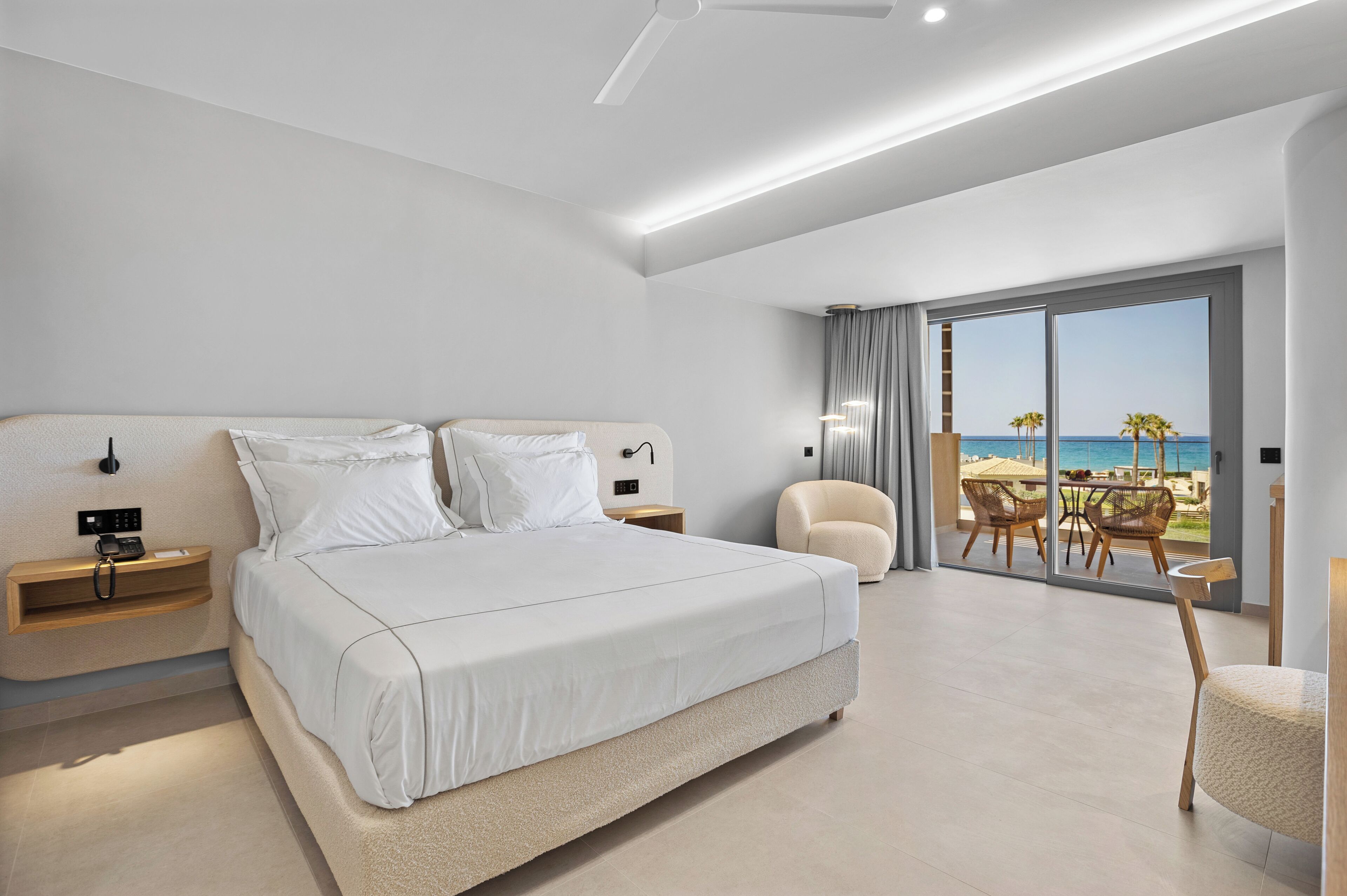 A modern hotel room with a large bed, bedside tables, a chair, and a balcony with a view of the sea.