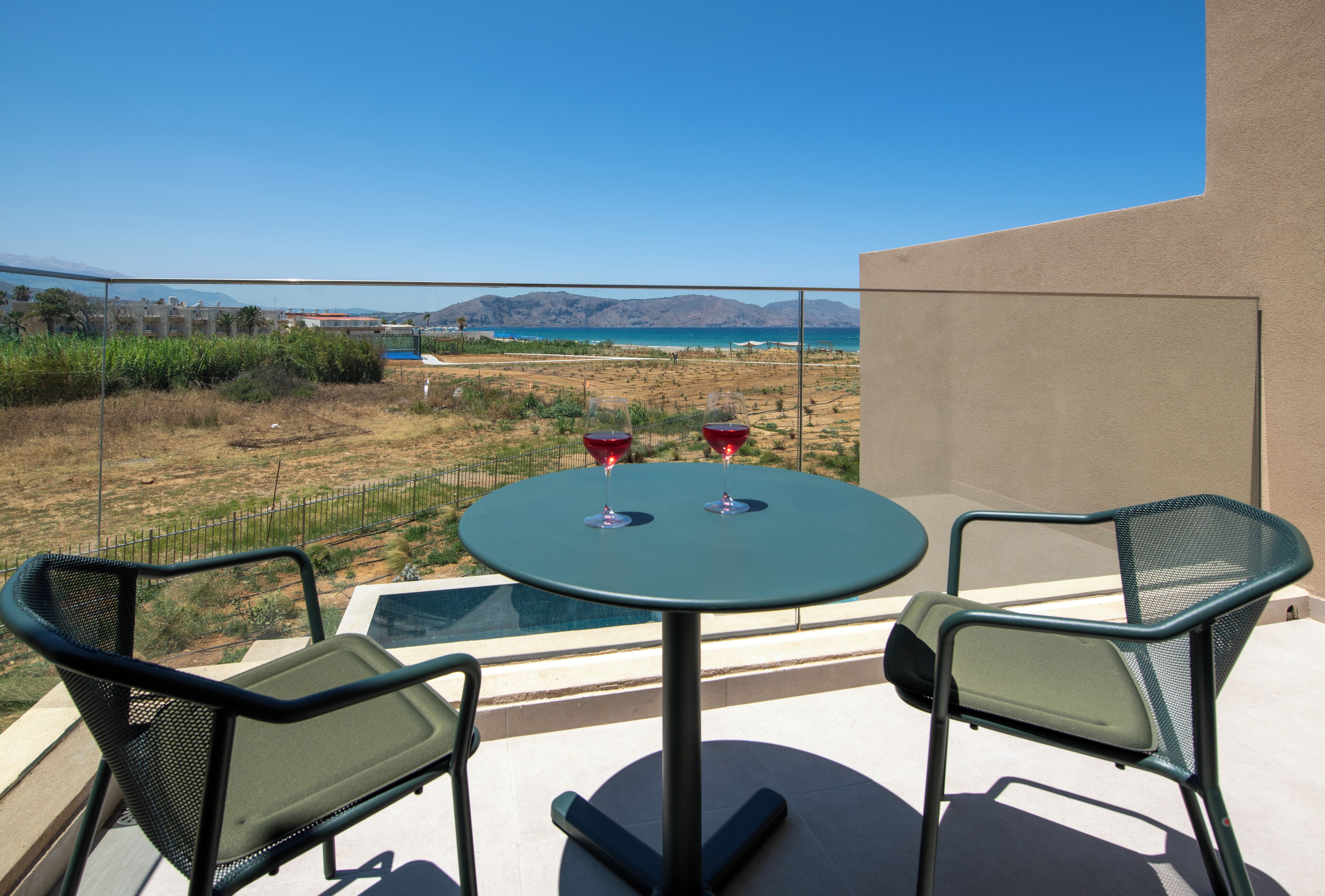 A balcony with a table and two chairs, overlooking a beach and mountains.