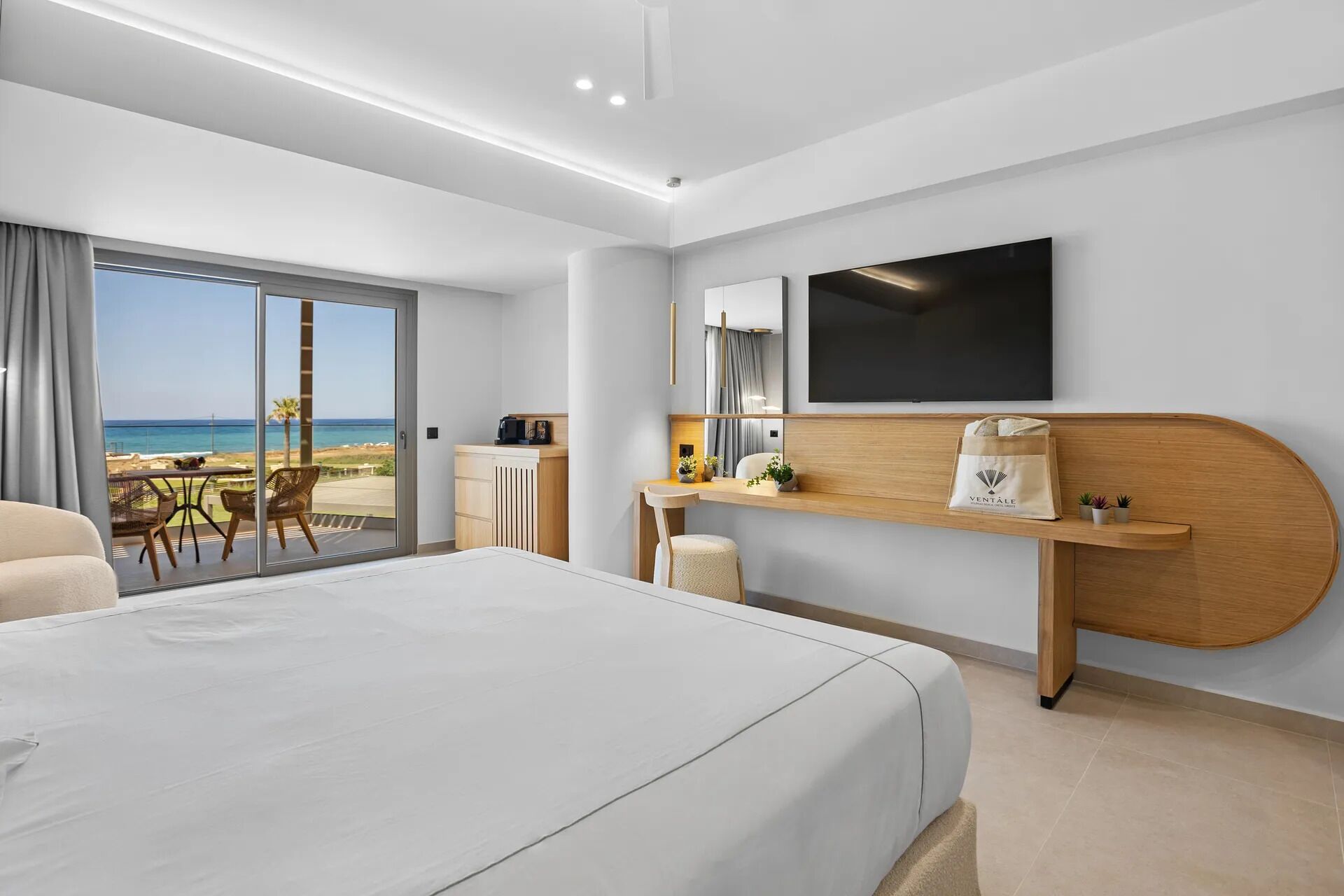 A modern hotel room with a large bed, a desk with a computer, a flat-screen TV, and a balcony with a view of the beach.