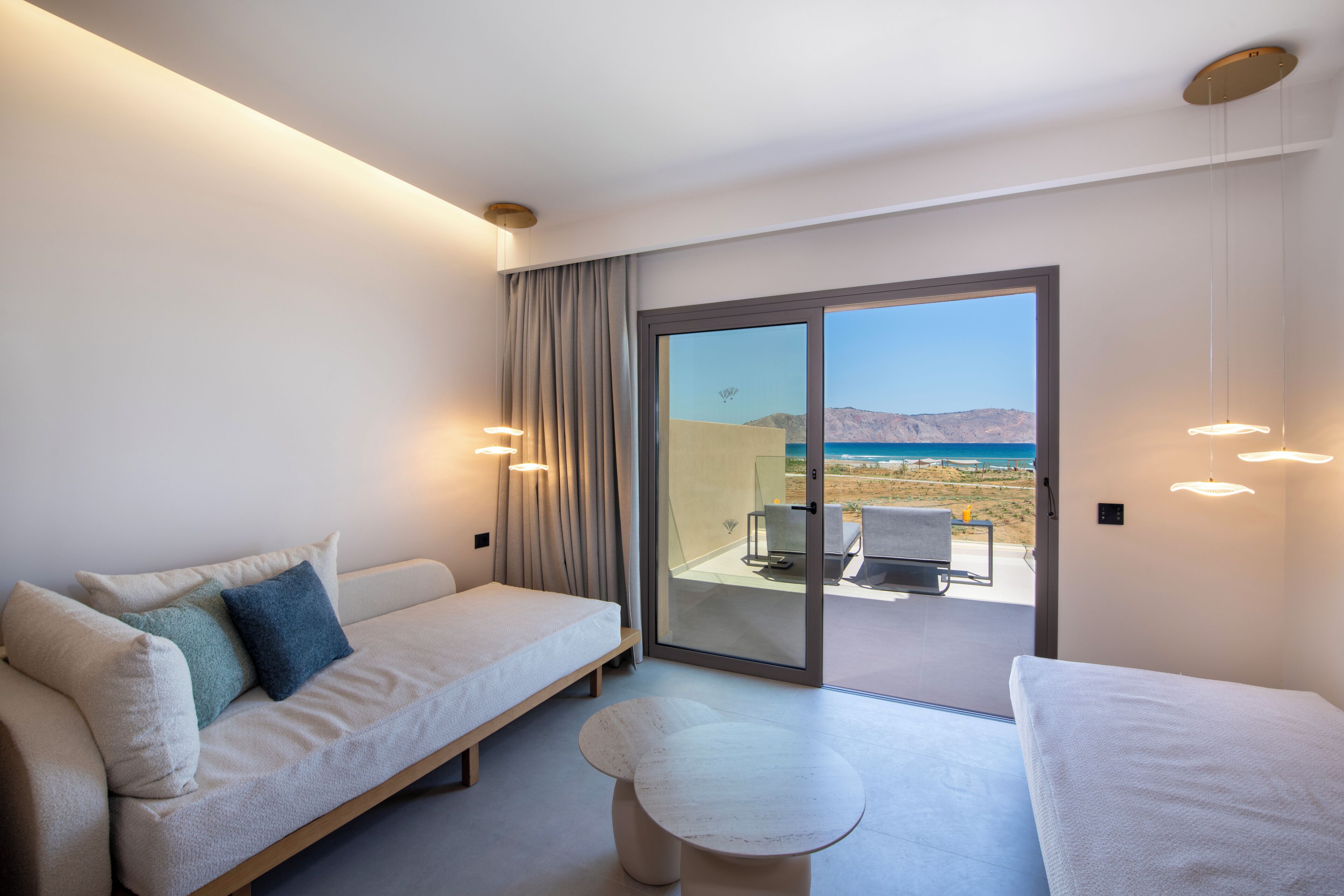 A modern hotel room with a sofa, a small round table, and a view of the beach through a sliding glass door.