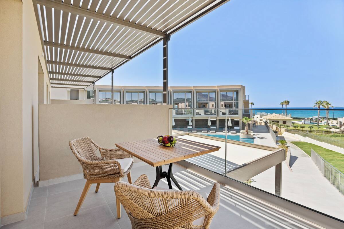 A balcony with wicker furniture and a table, overlooking a pool and a modern building.