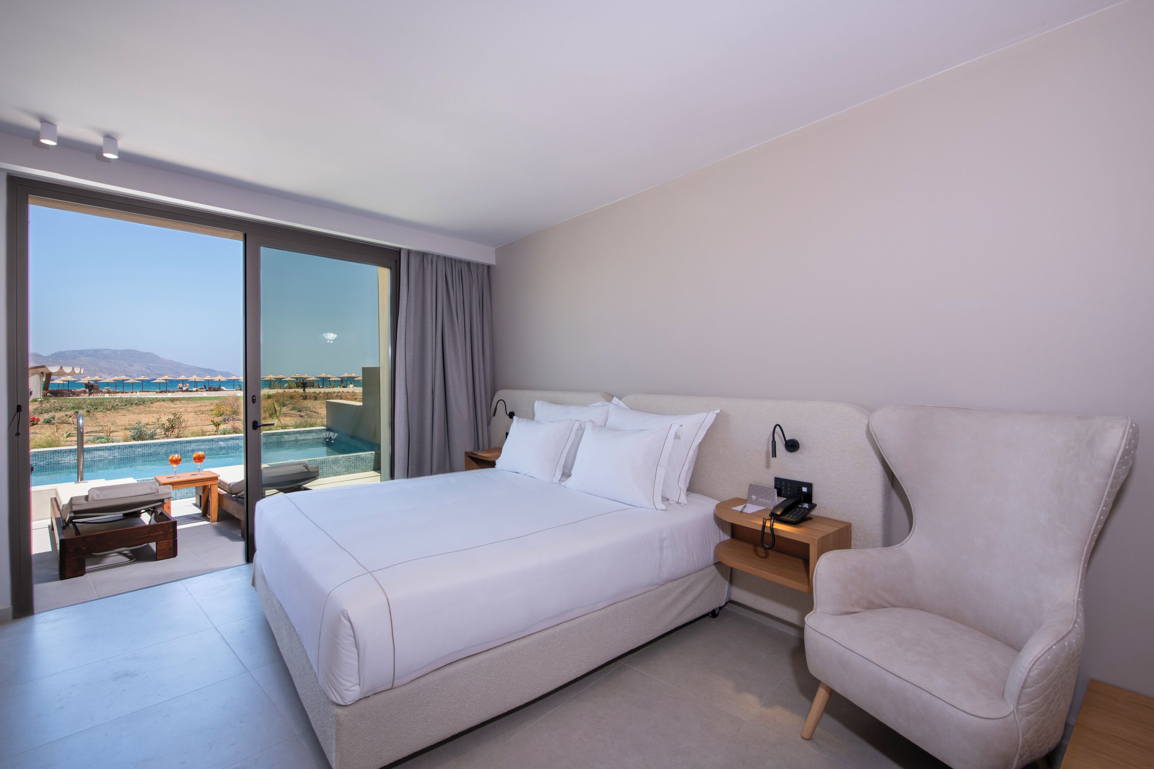 A modern hotel room with a large bed, a chair, a desk, and a view of a pool and mountains.