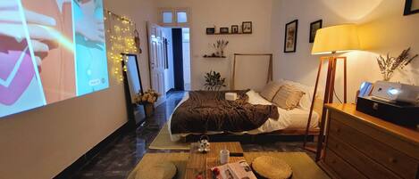 A cozy hotel room with a bed, bedside table, lamp, and a large screen displaying a movie.