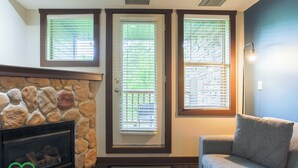 A living room with a stone fireplace, a couch, and two windows with blinds. - 2 BDRM Condo - Superb Facilities - Great Location (Fernie)