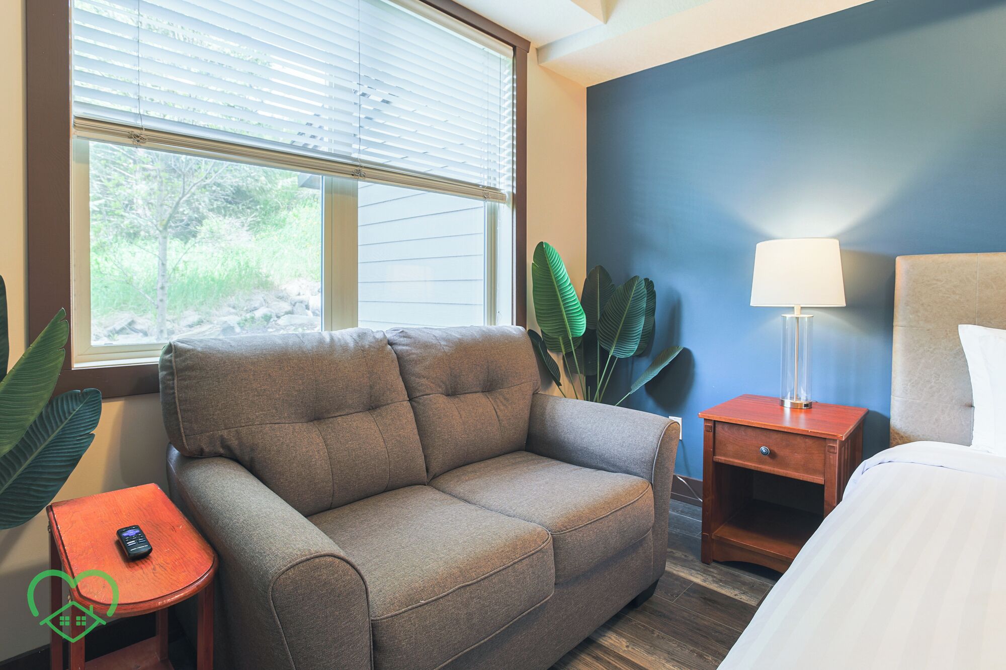 A room with a grey sofa, a small wooden side table with a lamp, a bed with white bedding, and a large window with blinds.